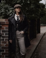 A person wearing a vintage-inspired outfit stands against a brick wall. The attire includes a newsboy cap, a tie, a waistcoat, and a long coat. The background is lush with greenery, giving a moody and classic feel.