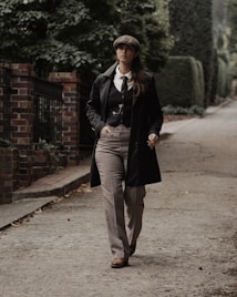 A person dressed in vintage-style clothing, including a flat cap and a long coat, walks down a tree-lined pathway. The path is bordered by a brick wall and lush greenery, creating a classic and timeless atmosphere.