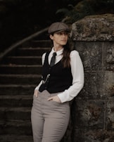A person in vintage attire stands thoughtfully against a moss-covered stone wall, wearing a flat cap, white dress shirt, vest, and checkered trousers. The background features dimly lit stone steps with foliage.
