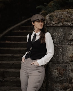 A person in vintage attire stands thoughtfully against a moss-covered stone wall, wearing a flat cap, white dress shirt, vest, and checkered trousers. The background features dimly lit stone steps with foliage.