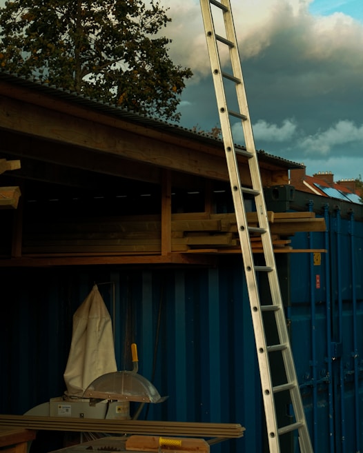 A metallic ladder is leaning against a wooden structure with stacks of wooden planks. There is a tree in the background under a cloudy sky. The structure is adjacent to a blue shipping container. Several tools and a covered saw are visible in the foreground.
