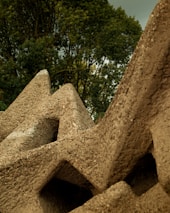 Large geometric concrete structures with rough surfaces are arranged closely together with dense green foliage in the background.