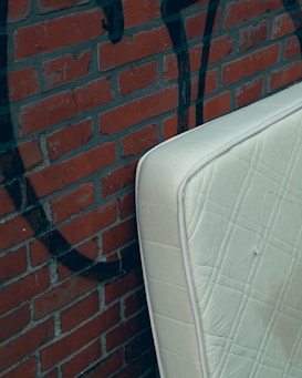 A light-colored mattress is leaning against a brick wall with graffiti. The wall is made of reddish-brown bricks with dark mortar lines. The graffiti features dark, abstract shapes.
