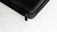 A close-up of a black textured hard case, featuring a zipper with a black pull tab. The background is a white surface with a subtle wood grain pattern.