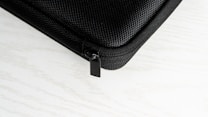 A close-up of a black textured hard case, featuring a zipper with a black pull tab. The background is a white surface with a subtle wood grain pattern.