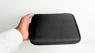 A hand holding a black, textured, rectangular case, likely made of fabric or synthetic material, against a light-colored background. The case appears to be a storage or carrying pouch.