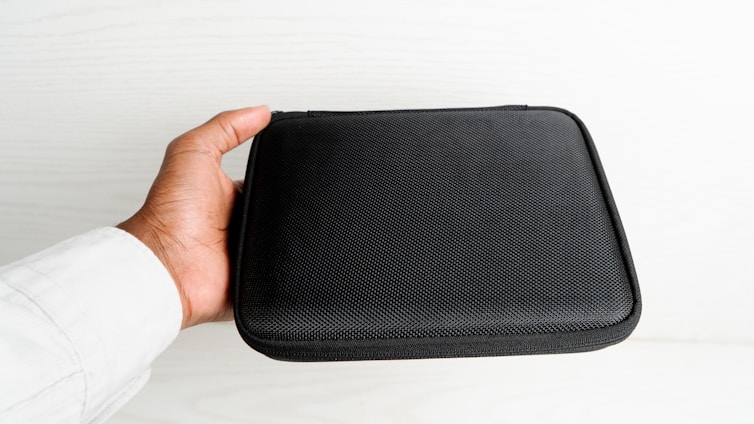 A hand holding a black, textured, rectangular case, likely made of fabric or synthetic material, against a light-colored background. The case appears to be a storage or carrying pouch.