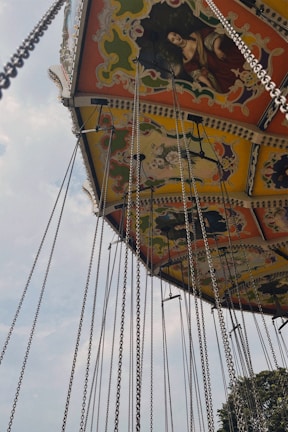 An ornate amusement park swing ride is depicted with colorful patterns and chains hanging down. The ride features painted panels with classical artwork. The sky is partially cloudy, adding to the vintage feel of the scene.