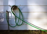 A green garden hose is looped and hanging on a wall-mounted holder against a white paneled wall. The hose extends down to a metal faucet situated on the left side of the image. Shadows of the hose and nearby foliage are cast on the wall, adding depth.