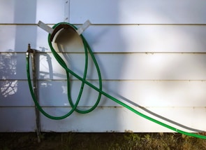 A green garden hose is looped and hanging on a wall-mounted holder against a white paneled wall. The hose extends down to a metal faucet situated on the left side of the image. Shadows of the hose and nearby foliage are cast on the wall, adding depth.