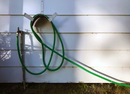 A green garden hose is looped and hanging on a wall-mounted holder against a white paneled wall. The hose extends down to a metal faucet situated on the left side of the image. Shadows of the hose and nearby foliage are cast on the wall, adding depth.