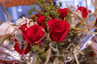 A bouquet of vibrant red roses and green leaves intermixed with small white flowers. The roses are central and prominent, with their petals beautifully unfurled. The green leaves and stems add a fresh contrast to the bright red roses, while white flowers provide delicate accents.