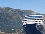 A large cruise ship is docked in front of a mountainous landscape. The ship has a prominent blue and white color scheme with multiple decks and windows. In the background, there is a range of rocky mountains covered in patches of green vegetation. Small houses and buildings are nestled at the base of the mountains.