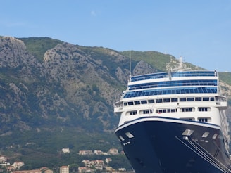 A large cruise ship is docked in front of a mountainous landscape. The ship has a prominent blue and white color scheme with multiple decks and windows. In the background, there is a range of rocky mountains covered in patches of green vegetation. Small houses and buildings are nestled at the base of the mountains.