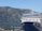A large cruise ship is docked in front of a mountainous landscape. The ship has a prominent blue and white color scheme with multiple decks and windows. In the background, there is a range of rocky mountains covered in patches of green vegetation. Small houses and buildings are nestled at the base of the mountains.