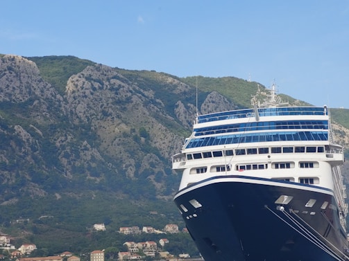 A large cruise ship is docked in front of a mountainous landscape. The ship has a prominent blue and white color scheme with multiple decks and windows. In the background, there is a range of rocky mountains covered in patches of green vegetation. Small houses and buildings are nestled at the base of the mountains.