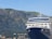 A large cruise ship is docked in front of a mountainous landscape. The ship has a prominent blue and white color scheme with multiple decks and windows. In the background, there is a range of rocky mountains covered in patches of green vegetation. Small houses and buildings are nestled at the base of the mountains.