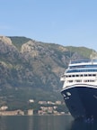 A large cruise ship partially visible on the right side, docked in calm waters with a backdrop of lush, rocky hills. Several small houses are nestled at the base of the hills.