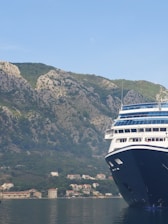 A large cruise ship partially visible on the right side, docked in calm waters with a backdrop of lush, rocky hills. Several small houses are nestled at the base of the hills.