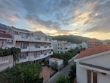 A residential area with multiple apartment buildings featuring red-tiled roofs and balconies. The view includes a street lined with lush green vegetation and a backdrop of tree-covered hills under a sky with dramatic cloud formations at sunset.