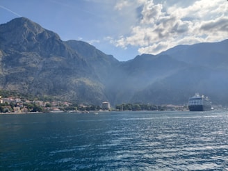 A large cruise ship sails near a mountain range, with a coastal town visible at the water's edge. Clear blue sky with some clouds adds to the scenic beauty of the area.
