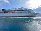 A large cruise ship is sailing on calm waters with a backdrop of mountains under a blue sky with scattered clouds. The ship is white with multiple decks and lifeboats lined along its side.