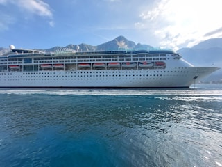 A large cruise ship is sailing on calm waters with a backdrop of mountains under a blue sky with scattered clouds. The ship is white with multiple decks and lifeboats lined along its side.