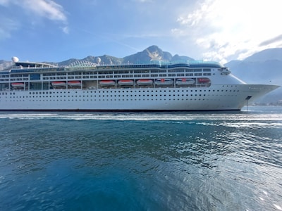 A large cruise ship is sailing on calm waters with a backdrop of mountains under a blue sky with scattered clouds. The ship is white with multiple decks and lifeboats lined along its side.
