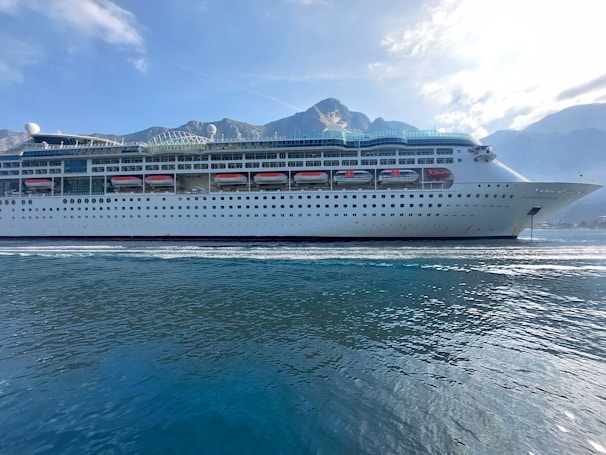 A large cruise ship is sailing on calm waters with a backdrop of mountains under a blue sky with scattered clouds. The ship is white with multiple decks and lifeboats lined along its side.