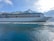 A large cruise ship is sailing on calm waters with a backdrop of mountains under a blue sky with scattered clouds. The ship is white with multiple decks and lifeboats lined along its side.
