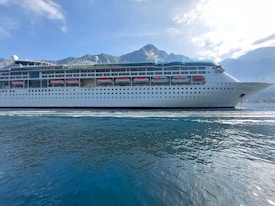 A large cruise ship is sailing on calm waters with a backdrop of mountains under a blue sky with scattered clouds. The ship is white with multiple decks and lifeboats lined along its side.