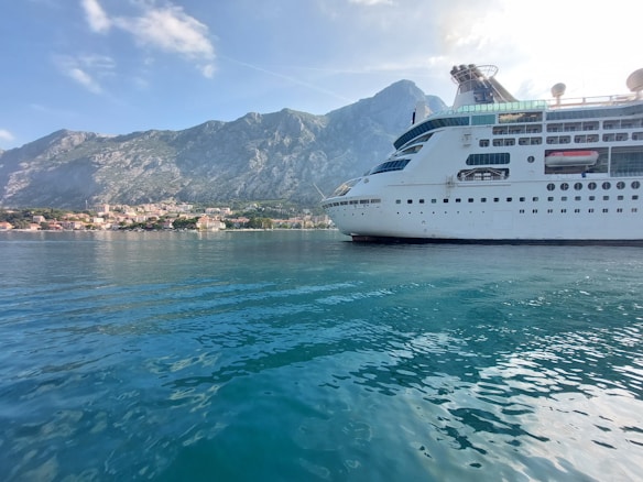 A large cruise ship is docked near a coastal town with a backdrop of majestic mountains. The water is a calm, clear blue, reflecting the ship and the sky above. The town is composed of clustered buildings with red-tiled roofs, nestled along the foot of the mountains under a partly cloudy sky.