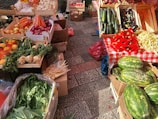 A vibrant market stall displays a variety of fresh produce. There are wooden crates filled with vegetables and fruits including carrots, red onions, zucchini, eggplants, red peppers, and watermelons. The stall features a red and white checkered tablecloth, set on a paved stone surface.