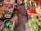 A vibrant market stall displays a variety of fresh produce. There are wooden crates filled with vegetables and fruits including carrots, red onions, zucchini, eggplants, red peppers, and watermelons. The stall features a red and white checkered tablecloth, set on a paved stone surface.