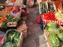 A vibrant market stall displays a variety of fresh produce. There are wooden crates filled with vegetables and fruits including carrots, red onions, zucchini, eggplants, red peppers, and watermelons. The stall features a red and white checkered tablecloth, set on a paved stone surface.