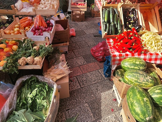 A vibrant market stall displays a variety of fresh produce. There are wooden crates filled with vegetables and fruits including carrots, red onions, zucchini, eggplants, red peppers, and watermelons. The stall features a red and white checkered tablecloth, set on a paved stone surface.