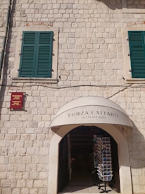 A stone building facade with two green shuttered windows and an arched entrance to a souvenir shop. A display stand with postcards is visible just inside the entrance. Above the entrance, there's a sign reading 'Forza Cattaro Souvenir Shop'. A small red plaque is mounted on the wall.