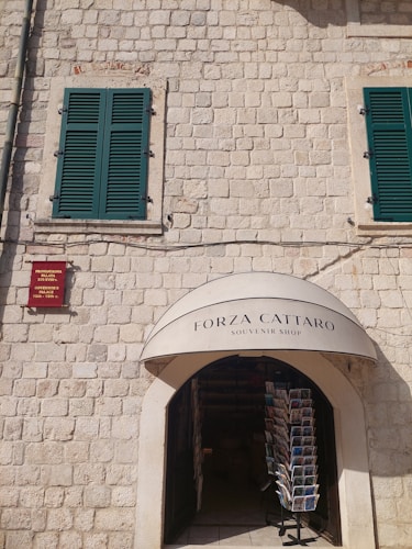 A stone building facade with two green shuttered windows and an arched entrance to a souvenir shop. A display stand with postcards is visible just inside the entrance. Above the entrance, there's a sign reading 'Forza Cattaro Souvenir Shop'. A small red plaque is mounted on the wall.