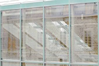 The image features a modern architectural facade composed of large glass panels. Through the glass, reflections of stone walls and window structures can be seen, creating a layered and abstract visual effect.