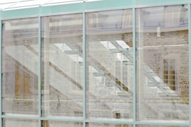 The image features a modern architectural facade composed of large glass panels. Through the glass, reflections of stone walls and window structures can be seen, creating a layered and abstract visual effect.