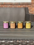 Four scented candles in jars are arranged in a row on a wooden table. Each jar is labeled with a different fragrance, including Fresh Mahogany, Sweet Pumpkin Pie, Amber, and Cedar And Saffron. The background features a wicker sofa and a brick wall.