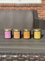 Four scented candles in jars are arranged in a row on a wooden table. Each jar is labeled with a different fragrance, including Fresh Mahogany, Sweet Pumpkin Pie, Amber, and Cedar And Saffron. The background features a wicker sofa and a brick wall.