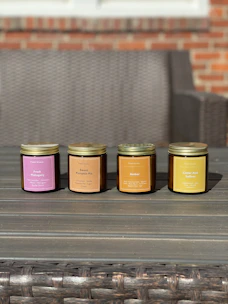 Four scented candles in jars are arranged in a row on a wooden table. Each jar is labeled with a different fragrance, including Fresh Mahogany, Sweet Pumpkin Pie, Amber, and Cedar And Saffron. The background features a wicker sofa and a brick wall.