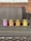 Four scented candles in jars are arranged in a row on a wooden table. Each jar is labeled with a different fragrance, including Fresh Mahogany, Sweet Pumpkin Pie, Amber, and Cedar And Saffron. The background features a wicker sofa and a brick wall.