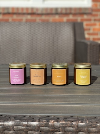 Four scented candles in jars are arranged in a row on a wooden table. Each jar is labeled with a different fragrance, including Fresh Mahogany, Sweet Pumpkin Pie, Amber, and Cedar And Saffron. The background features a wicker sofa and a brick wall.