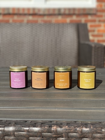 Four scented candles in jars are arranged in a row on a wooden table. Each jar is labeled with a different fragrance, including Fresh Mahogany, Sweet Pumpkin Pie, Amber, and Cedar And Saffron. The background features a wicker sofa and a brick wall.