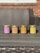 Four scented candles in jars are arranged in a row on a wooden table. Each jar is labeled with a different fragrance, including Fresh Mahogany, Sweet Pumpkin Pie, Amber, and Cedar And Saffron. The background features a wicker sofa and a brick wall.