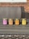 Four scented candles in jars are arranged in a row on a wooden table. Each jar is labeled with a different fragrance, including Fresh Mahogany, Sweet Pumpkin Pie, Amber, and Cedar And Saffron. The background features a wicker sofa and a brick wall.