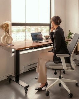 A person sitting in an ergonomic chair at a modern desk by a large window, looking thoughtfully outside. The desk features a closed laptop, a decorative vase with dried plants, and a small bowl. The room is well-lit and has a minimalist, cozy atmosphere.