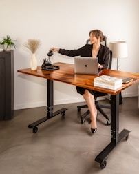 A woman in a black suit is seated at a wooden desk with an open laptop in front of her. She is reaching for a traditional black rotary phone on the desk. The workspace includes decorative elements such as a vase with dried grass, a small plant, and a stack of books. A lamp stands in the background.